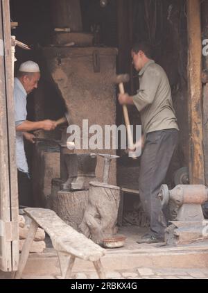 Two blacksmiths working in roadside workshop in Kashgar markets. One working at furnace and the other using sledgehammer. Clay furnace in centre. Stock Photo