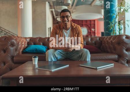 Smiling male freelancer sitting on sofa and looking at camera during working day in coworking Stock Photo