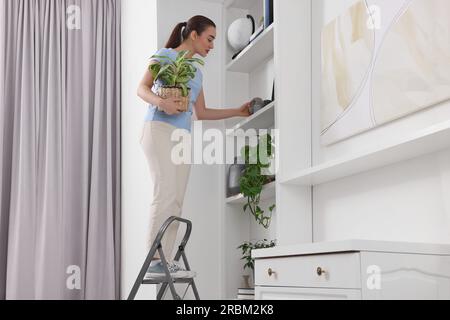 Woman on ladder with houseplant near shelves at home Stock Photo