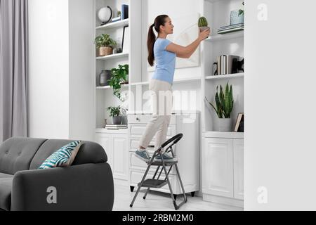 Woman on ladder with houseplant near shelves at home Stock Photo