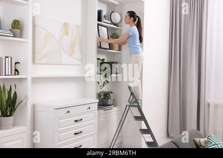 Woman on ladder with picture near shelves at home Stock Photo