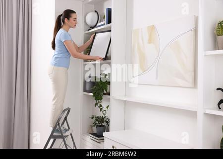 Woman on ladder with picture near shelves at home Stock Photo