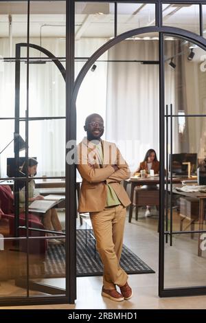 Young confident African American businessman in beige formalwear looking at camera while standing in arch shaped door of coworking area Stock Photo
