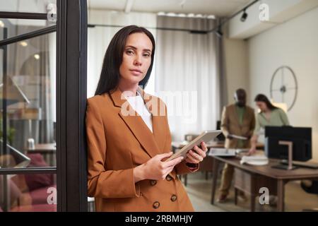 Young serious businesswoman in formalwear holding tablet and looking at you while standing by open door of coworking area with her colleagues Stock Photo