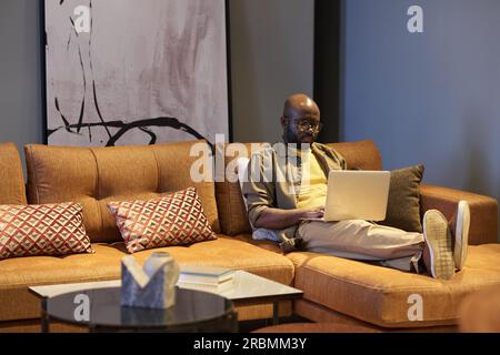 Serious young black man with laptop on his knees sitting on comfortable couch with cushions in living room while working over new project Stock Photo