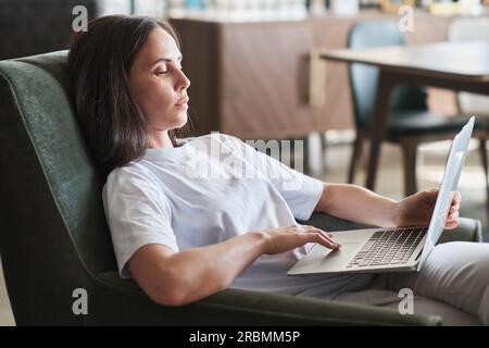 Young serious businesswoman with laptop sitting in comfortable armchair and looking at screen while watching online video or reading data Stock Photo