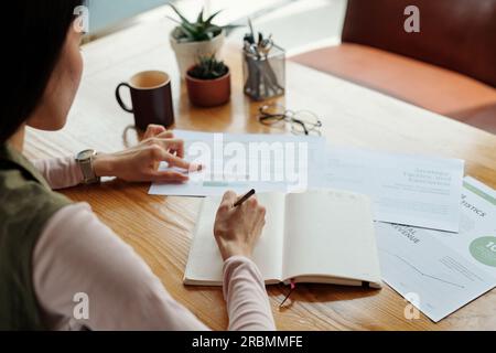 Close-up of young businesswoman with pen over blank page of open notebook making notes while analyzing financial data in document Stock Photo