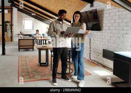 Long shot of two young intercultural managers in casualwear looking at laptop screen while African American man explaining online data Stock Photo