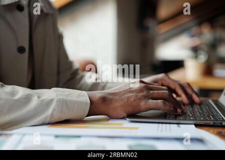 Focus on hands of young African American male broker typing on laptop keyboard while sitting by workplace with financial documents Stock Photo