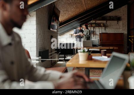 Focus on two young analysts or managers looking at screen of tablet held by brunette woman and discussing online data in loft coworking space Stock Photo
