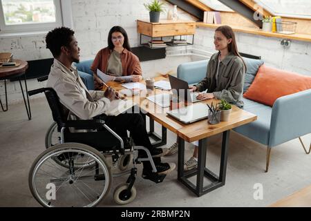 Two young intercultural female managers looking at confident African American male colleague in wheelchair during discussion at meeting Stock Photo