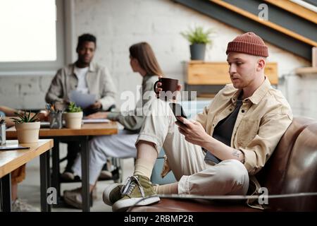 Modern businessman in casualwear having coffee and texting in mobile phone at break while sitting on leather couch against colleagues Stock Photo