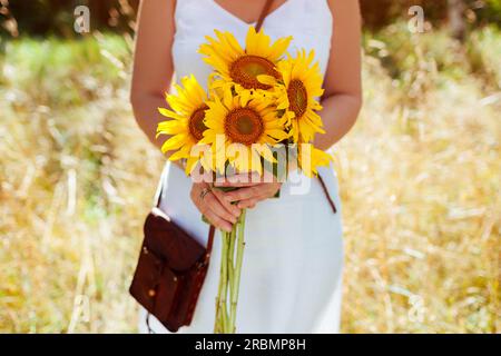 Woman holding sunflower bouquet outdoors wearing white dress holding handbag in meadow. Summer boho rustic style Stock Photo