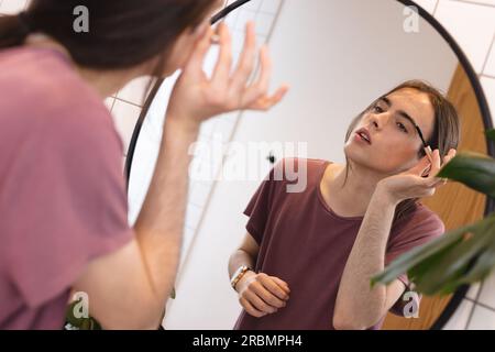Non-binary transgender woman applying eyebrow makeup looking in bathroom mirror at home. Gender fluidity, health, beauty and lifestyle. Stock Photo