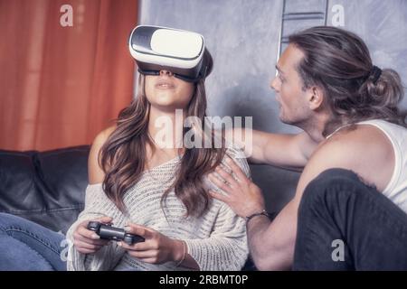 Cheerful modern couple of caucasian guys play together in living room of their studio house. Male guy enjoyng life with a girl playng videogames with Stock Photo