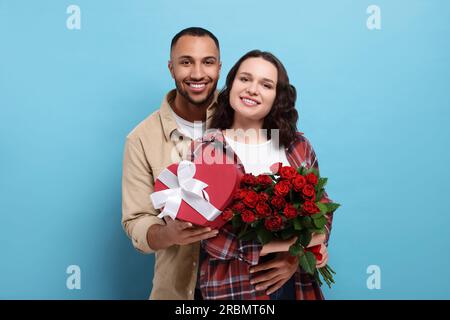 Lovely couple with gift box and bouquet of red roses on light blue background. Valentine's day celebration Stock Photo