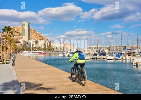 nice senior woman cycling on a cool spring day with her electric bicycle at the waterfront of Alicante, Costa Blanca, Spain Stock Photo