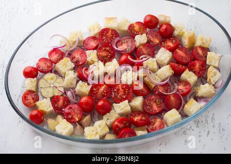 Cherry tomatoes, croutons, red onion, garlic and spices in a glass baking dish on a light gray background, top view. cooking italian panzanella salad Stock Photo