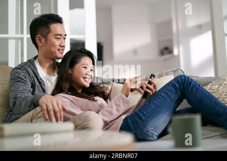 happy young asian couple sitting on family couch at home looking at cellphone photos together Stock Photo