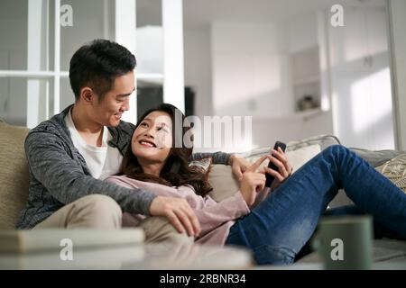 happy young asian couple sitting on family couch at home looking at cellphone photos together Stock Photo