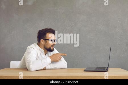 Happy young man sitting at a desk and pointing at the screen of a laptop computer Stock Photo