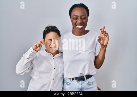 Young mother and son standing together over white background gesturing finger crossed smiling with hope and eyes closed. luck and superstitious concep Stock Photo