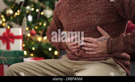 Young bald man sitting by christmas tree suffering stomachache at home Stock Photo