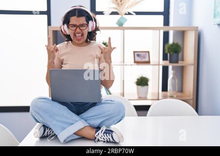 Young hispanic woman using laptop sitting on the table wearing headphones shouting with crazy expression doing rock symbol with hands up. music star. Stock Photo