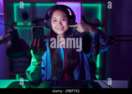 Young asian woman playing video games with smartphone smiling cheerful showing and pointing with fingers teeth and mouth. dental health concept. Stock Photo