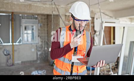 Young hispanic man builder talking on walkie talkie using laptop at construction site Stock Photo
