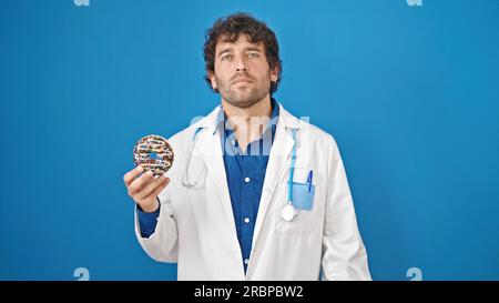 Young hispanic man nutritionist holding doughnut with serious face over isolated blue background Stock Photo
