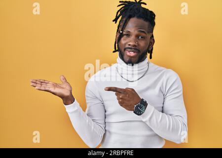 African man with dreadlocks wearing turtleneck sweater over yellow background amazed and smiling to the camera while presenting with hand and pointing Stock Photo