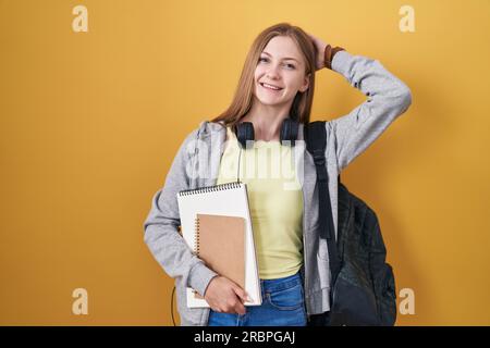 Young caucasian woman wearing student backpack and holding books smiling confident touching hair with hand up gesture, posing attractive and fashionab Stock Photo