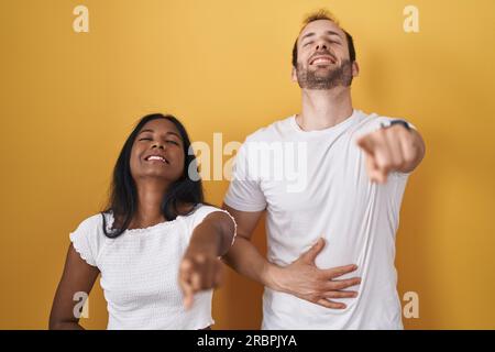 Interracial couple standing over yellow background laughing at you, pointing finger to the camera with hand over body, shame expression Stock Photo