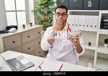 Young hispanic doctor man with beard holding candy covering mouth with hand, shocked and afraid for mistake. surprised expression Stock Photo