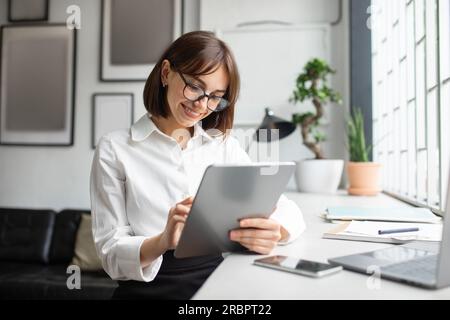 Confident european businesswoman working online, typing on tablet for investment or chatting with clients in office Stock Photo