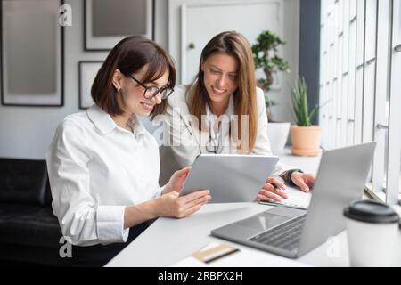 Two lady colleagues discussing new project, businesswomen using digital tablet together in office Stock Photo