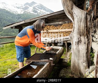 A gray-haired man in sunglasses washes his hands at a village spring against the backdrop of a firewood shed, Austria Stock Photo