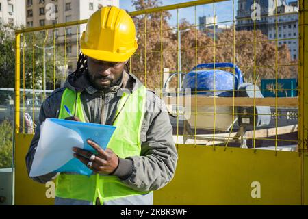 young black man of african ethnicity, wearing green vest jacket and yellow safety helmet, construction worker at his outdoor workspace making notes in Stock Photo