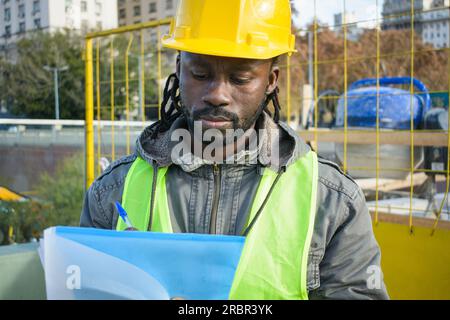 closeup of young African engineer man with dreadlocks and beard, wears vest and safety helmet you are standing at his work focused taking notes in a f Stock Photo