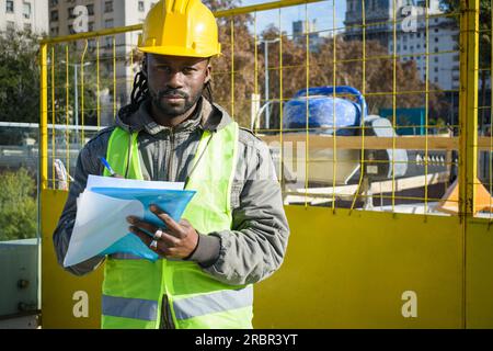 Black male engineer with beard and dreadlocks, wearing yellow safety helmet and green vest, standing outside on a construction site holding a folder i Stock Photo
