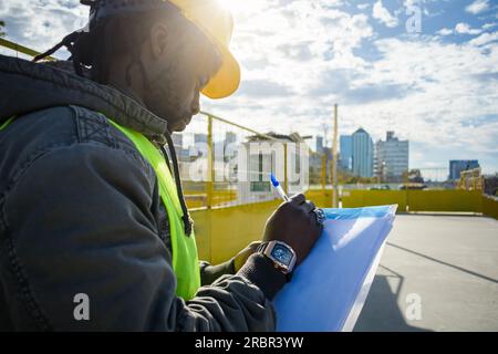 rear view of male African ethnicity engineer wearing hardhat and vest, standing outside at his workspace at sunset making notes on a paper, focus on h Stock Photo