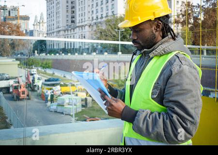 young black engineer man wearing yellow safety helmet and vest, standing outdoors supervising the construction site making notes in a folder, copy spa Stock Photo