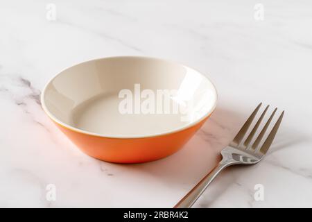 Orange bowl and metal fork over white marble table. Empty porcelain ramekin for food design mockup. Modern tableware and ceramic crockery concept. Stock Photo