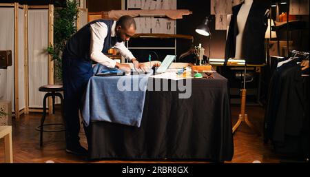 Young tailor marking fabric cloth to design fashion collection in tailoring workshop, using chalk for handcraft. Designer couturier working on luxury clothes creation, craftsmanship. Stock Photo