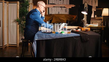 Old designer calling customer in atelier, discussing about measurements and fabric on phone call. Senior tailor working on custom made clothing line to create tailored items. Handheld shot. Stock Photo