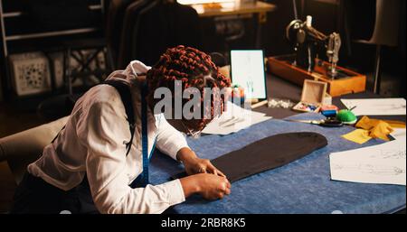 African american tailor preparing fabric to create tailored suit in atelier, working with crafting tools and materials for custom made designs. Young seamstress manufacturing handmade items. Stock Photo