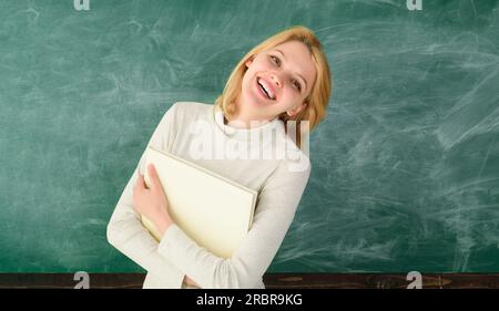 Portrait of female teacher in classroom front chalkboard. Smiling teacher with notebook near blackboard. Teachers day. Happy student, teacher in Stock Photo