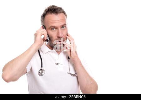 Middle-aged male doctor in a white shirt and stethoscope taking a call on his mobile phone with a worried expression as he listens to the Stock Photo