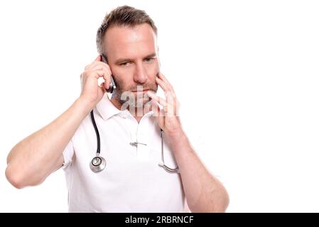 Middle-aged male doctor in a white shirt and stethoscope taking a call on his mobile phone with a worried expression as he listens to the Stock Photo